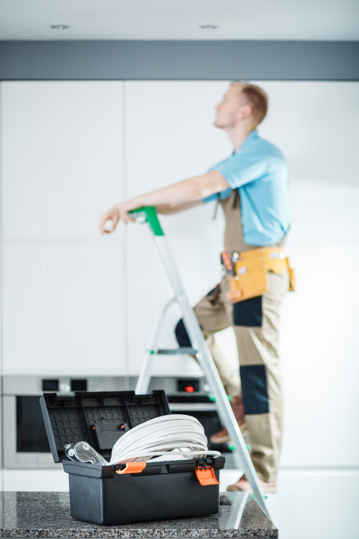 Black toolbox on the countertop and electrician on a ladder repairing lighting in the kitchen